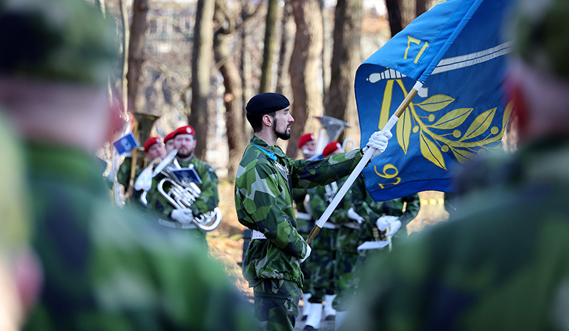 fokus på en militärklädd man med en flagga i handen. I bakgrunden ser man flera militärer med bläckblåsinstrument.