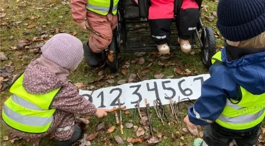Foto på barn som är ute i skogen, de sitter på marken och lägger 1 pinne på siffran 1, 2 pinnar på siffran 2 o s v.