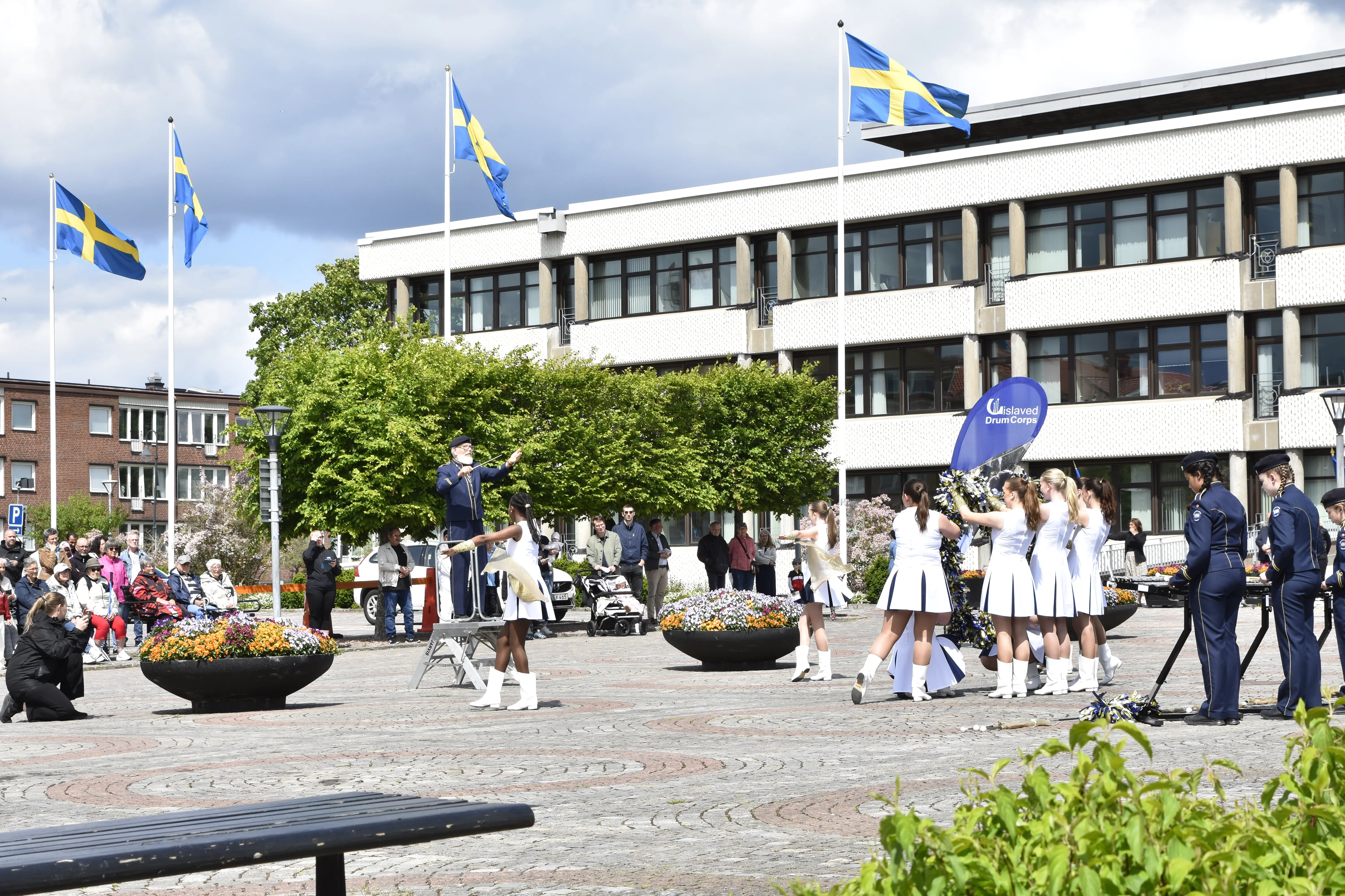 Ett torg med drillflickor och publik. I bakgrunden syns ett stort hus med fyra flaggstänger framför med Sverigeflaggan hissad.