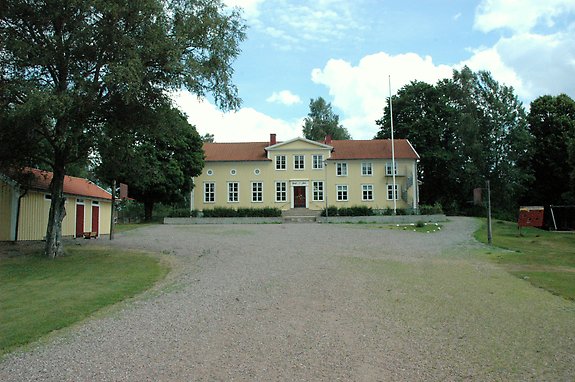 en äldre gul skola med en stor gårdsplan och en flaggstång till höger framför skolan.