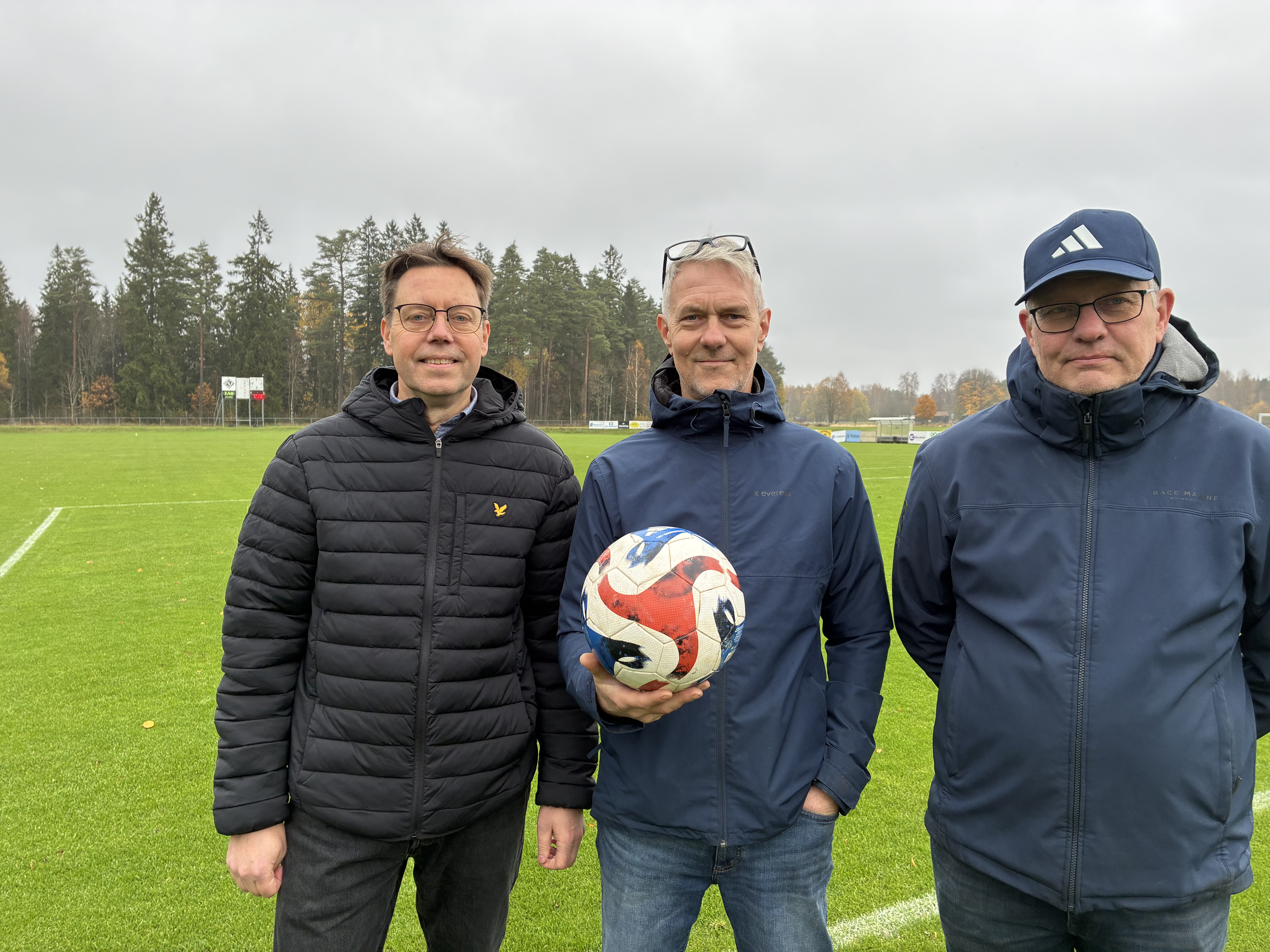 Tre personer från föreningen står framför en fotbollsplan och håller en fotboll.