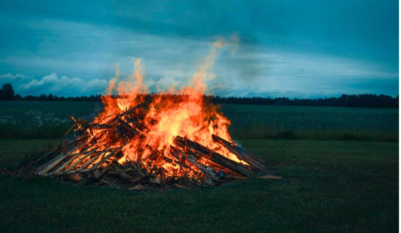 En stor brasa av virke brinner på en grön äng under en blå himmel.