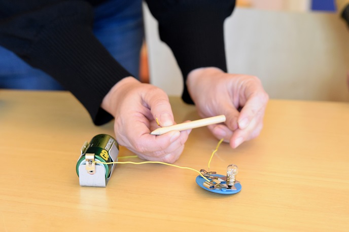 Ett par händer demonstrerar ett experiment med ett batteri, en blödlampa och en blyertspenna.