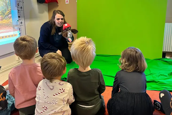 Man ser ryggar på barn som sitter framför green screen, Elida håller i handdockan och har samling med barnen.