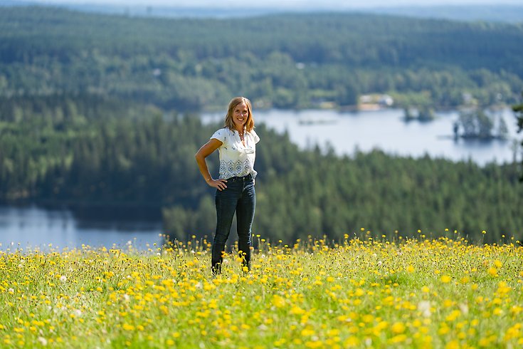 En kvinna står på en höjd med sjö och skog i bakgrunden.