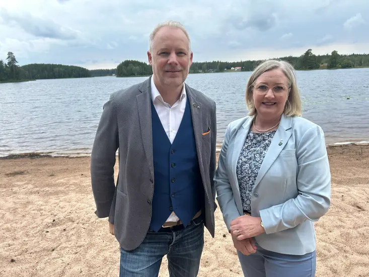 Anton Sjödell och Kristine Hästmark står vid en strand med en sjö i bakgrunden. 