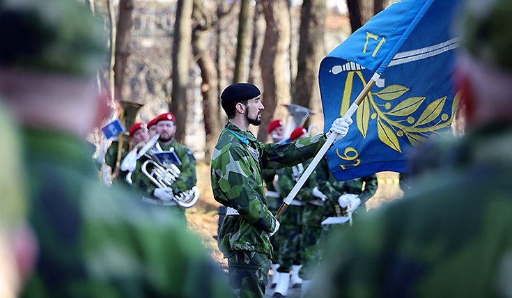 fokus på en militärklädd man med en flagga i handen. I bakgrunden ser man flera militärer med bläckblåsinstrument.