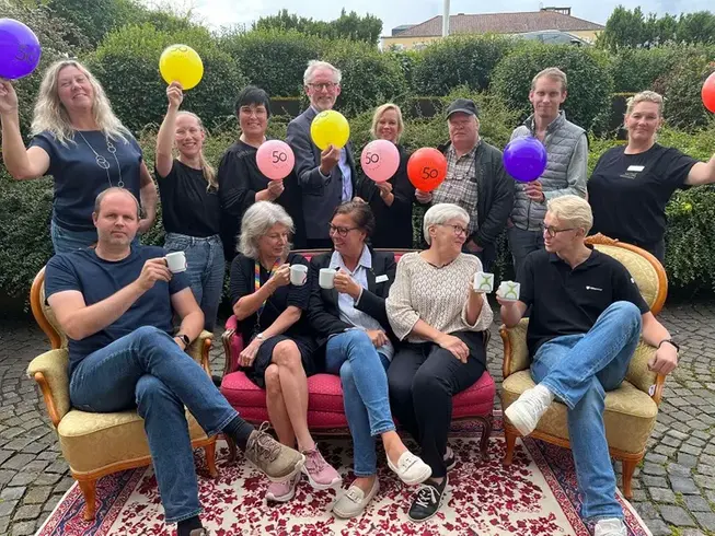 Tre kvinnor sitter i en rosa soffa med kaffekoppar omgiven av ett gäng män och kvinnor med ballonger