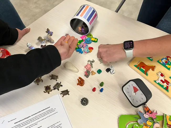 Foto på små leksaker som ligger på ett bord, händer som sorterar dem efter vad det är: tärningar, djur, suddin med mera.