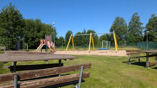 En lekplats med gungor och en rutschkana, samt träbord och bänkar på gräsmattan. Skylten på rutschkanan visar ”'Lekplats'".