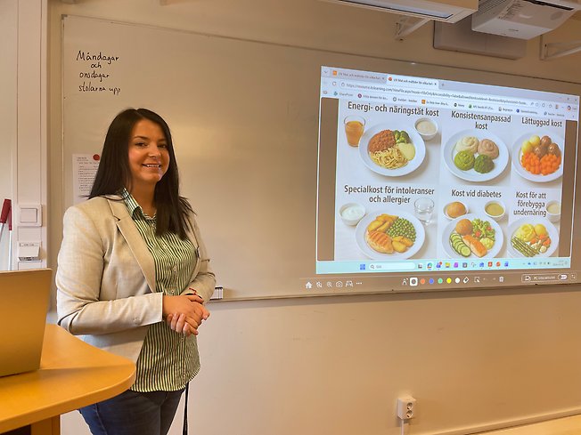 En kvinna står längst fram i ett klassrum framför en whiteboard.