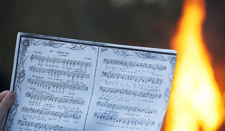 En hand håller i ett pappersark med noterna till låten "Sköna maj". I bakgrunden syns en eld.