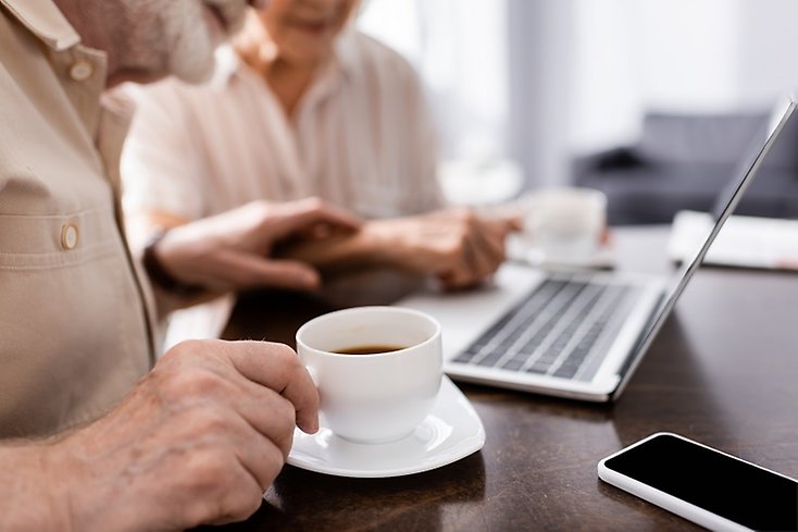 Dator och kaffekopp med två äldre personer i bakgrunden