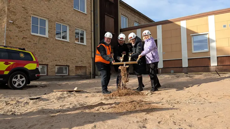 4 personer tar ett gemensamt spadtag i en hög av sand med hjälp av en specialbyggd spade med fyra handtag. De står på platsen där den nya Brandstationen ska föras upp.