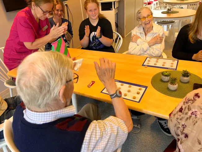 Foto på en äldre herre som lyfter armrna i triumf, alla runt bordet applåderar.