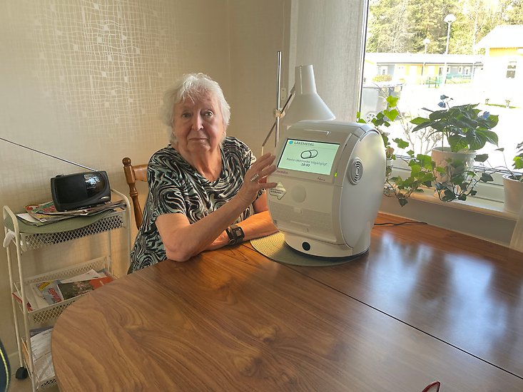 Äldre kvinna sitter vid sitt köksbord bredvid hennes medicingivare.