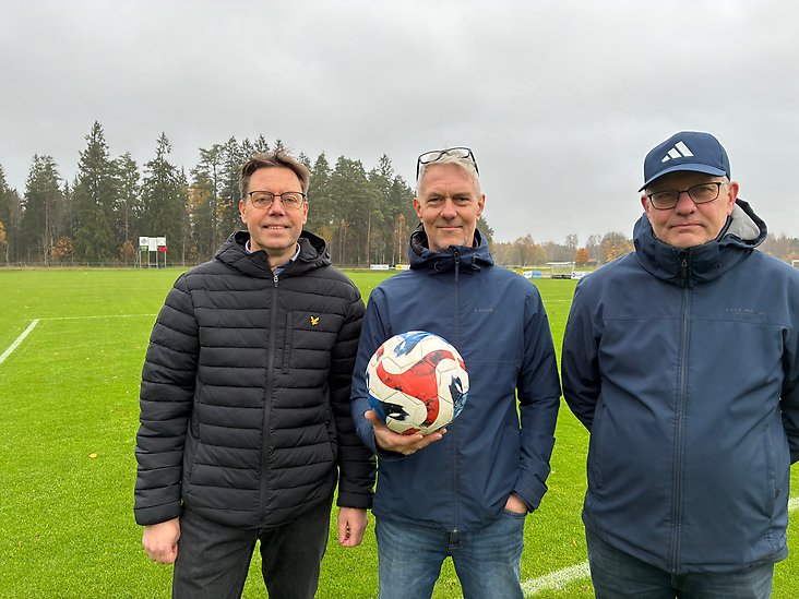 Tre personer från föreningen står framför en fotbollsplan och håller en fotboll.