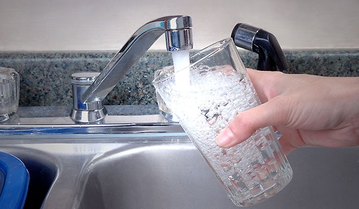 En hand som håller i ett vattenglas. Vattenglaset fylls med vatten från en kökskran